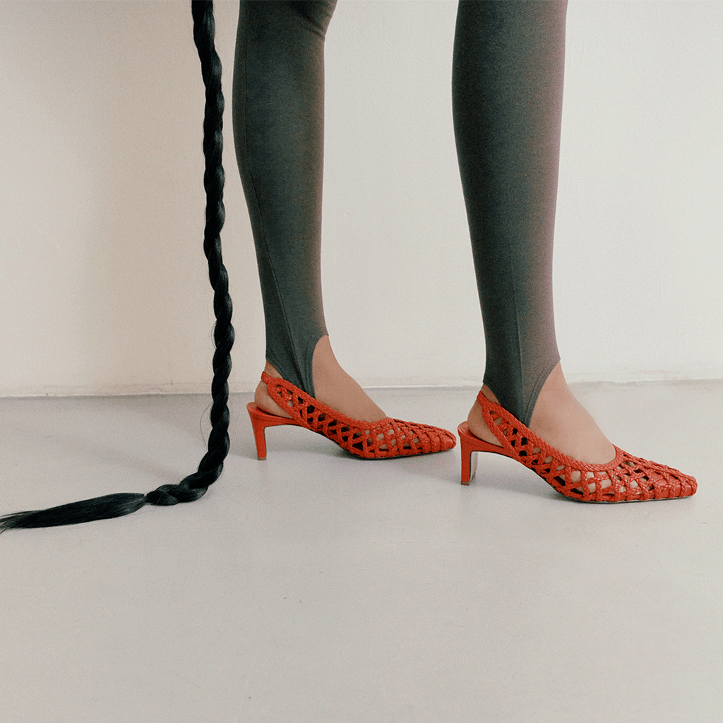 Cora braided leather heels in red, shown on both feet in motion, styled with a long braided hair prop and neutral tones.


