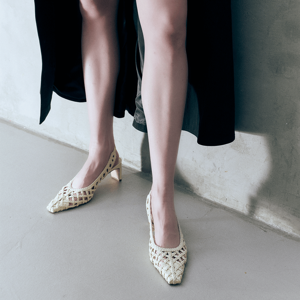 Model wearing Daniella Shevel's Cora braided leather slingback heels in Stone Meringue, styled with a black outfit to highlight the contrast and pointed silhouette.

