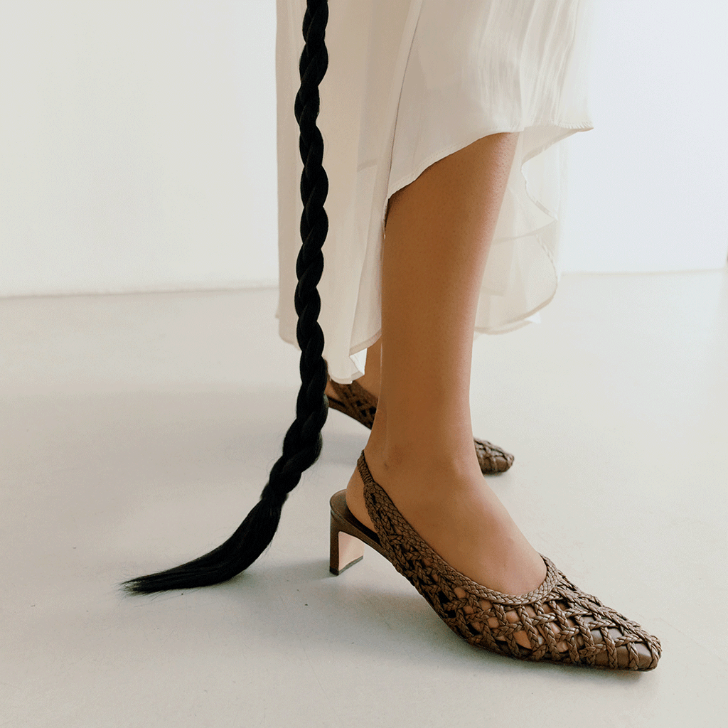 Profile view of the Daniella Shevel Cora braided slingback heels in brown worn with a white flowy dress, showcasing heel and texture.

