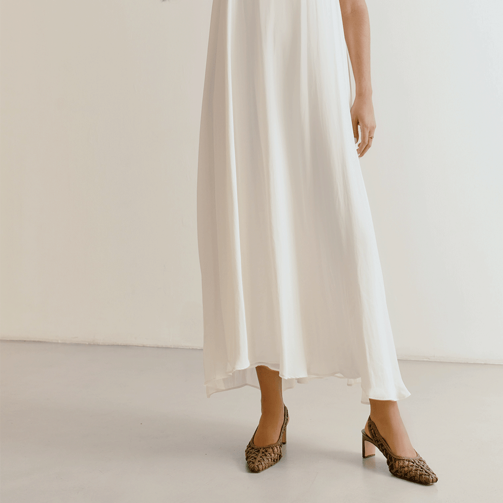 Model wearing the brown Cora braided leather slingback heels by Daniella Shevel with an ivory flowy dress, showcasing the elegant pointed toe and mid-heel style.

