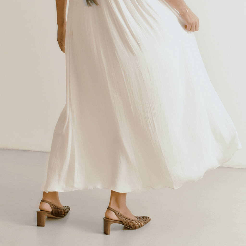 Back angle of Daniella Shevel’s Cora slingback heels in brown, featuring woven texture and block heel paired with an ivory dress.


