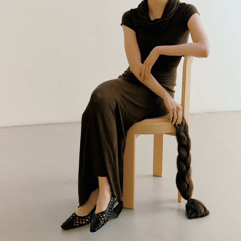 Full-body seated shot of model wearing Halo flats in Black – paired with a chocolate knit dress and relaxed elegance.