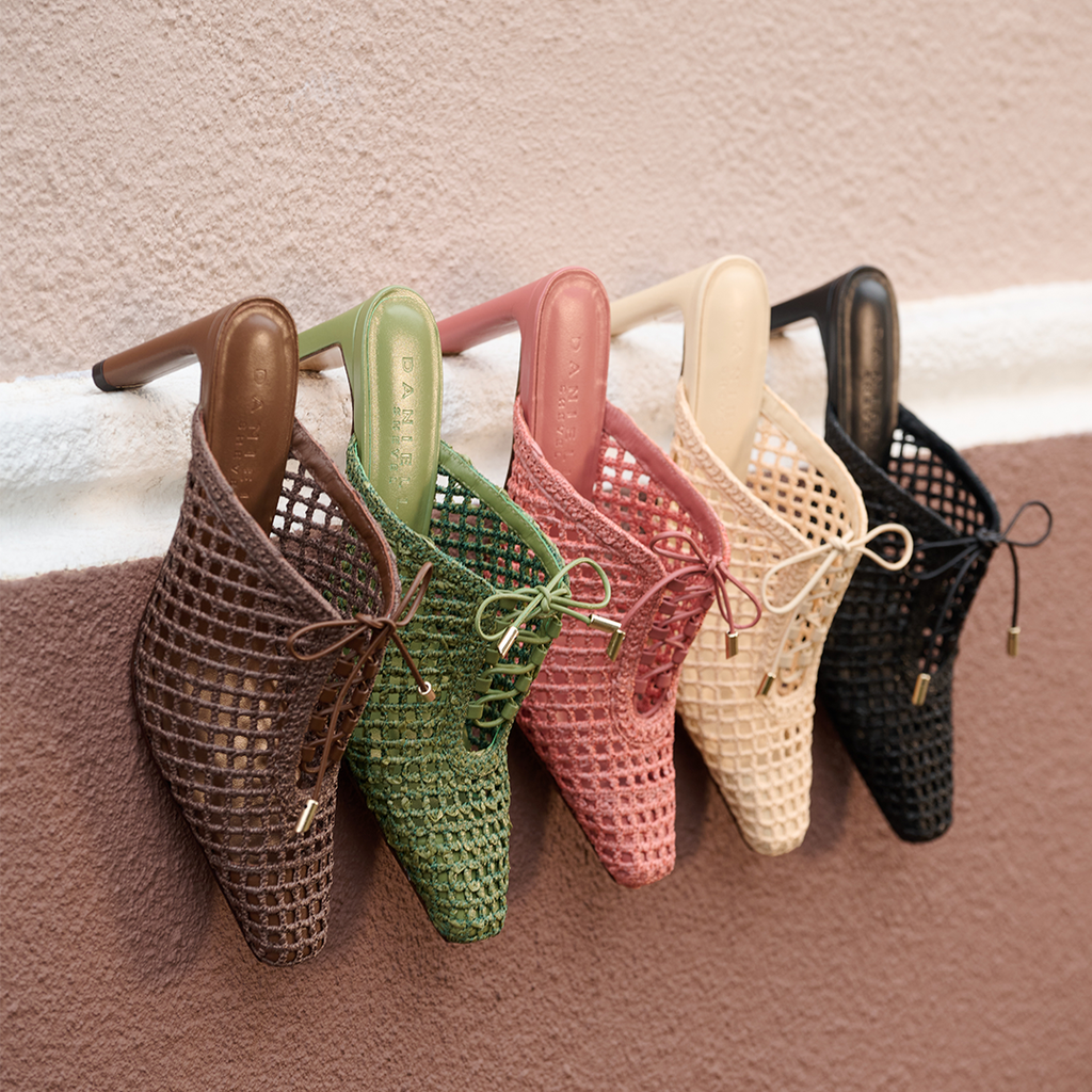 Daniella Shevel Lina raffia mules in a tonal range including Wild Straw, styled in a soft cascading lineup from a white railing for a fresh, summery mood.