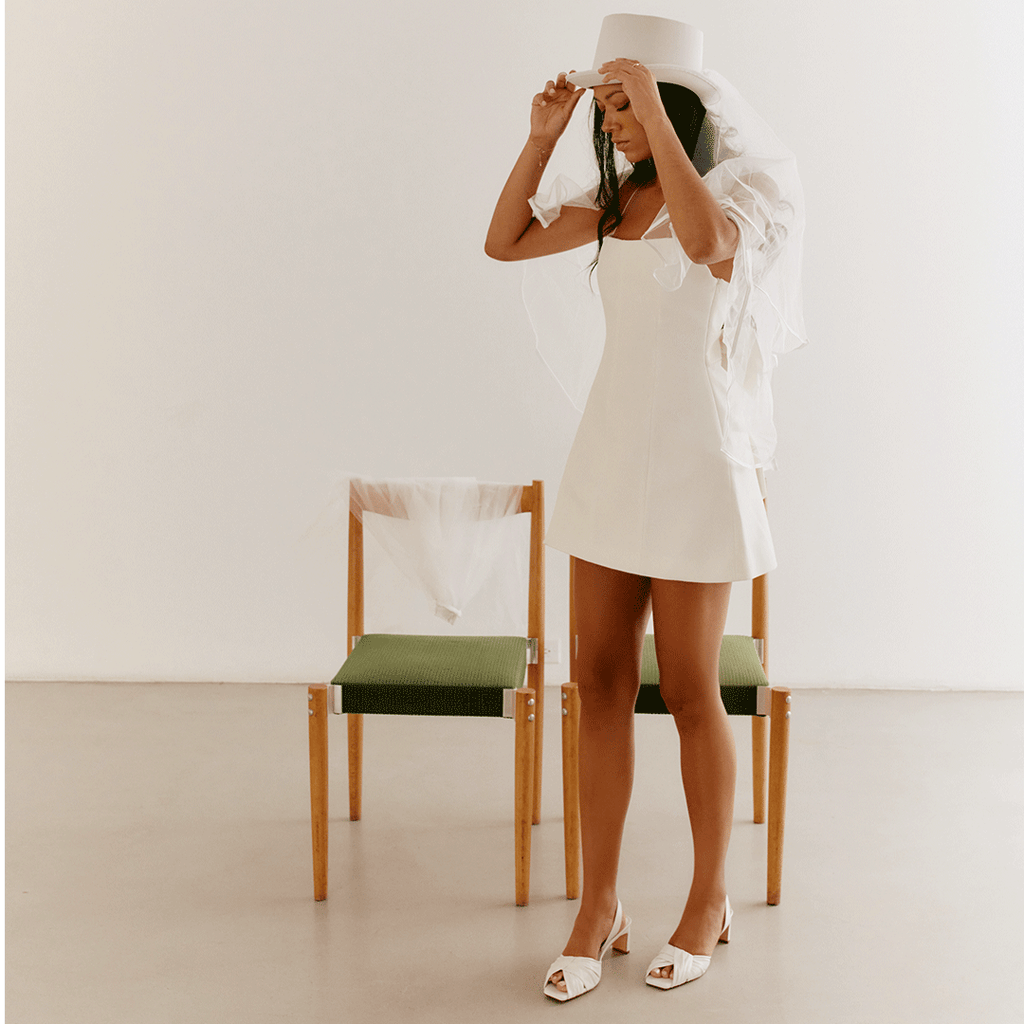 Model in white dress seated, showcasing Riley white low-heel sandals.