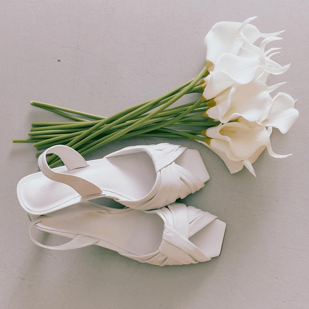 Overhead shot of Riley white sandals styled beside white lilies.