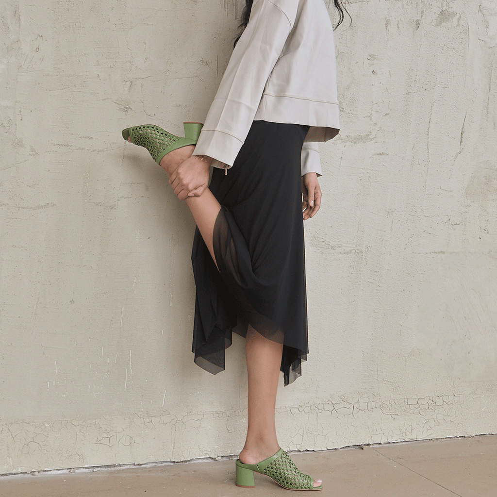 Close-up of a woman’s legs in Daniella Shevel green woven mule, highlighting movement and flow of a pleated skirt.